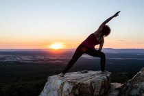 Junge Yogi-Frau praktiziert Yoga auf einem Felsen im Gebirge im Licht des Sonnenaufgangs, Vorderansicht mit einem Arm nach oben — Stockfoto