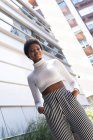 Dal basso elegante donna afroamericana sicura di sé con le mani sulla tasca guardando la fotocamera mentre in piedi vicino a moderni condomini nella giornata di sole in città — Foto stock