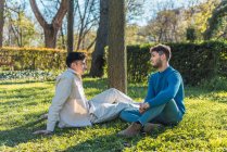 Vista lateral de casal sorridente de homens homossexuais sentados no gramado no parque e desfrutando de dia ensolarado enquanto olham um para o outro — Fotografia de Stock