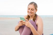 Delighted female in activewear sitting on yoga mat browsing on mobile phone on seashore — Stock Photo