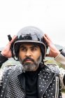 Самоуверенный взрослый бородатый велосипедист в стильной кожаной куртке, регулирующий защитный шлем и смотрящий в сторону в природе — стоковое фото