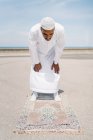 Menino completo macho islâmico em roupas brancas tradicionais em pé no tapete e orando contra o céu azul na praia — Fotografia de Stock