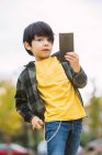 Adorable boy with black hair in casual clothes browsing cellphone and looking at camera while standing on city street on blurred background — Stock Photo