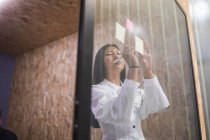 Sorrindo Asiático empregado feminino escrevendo na nota pegajosa na parede de vidro enquanto trabalhava no escritório moderno — Fotografia de Stock