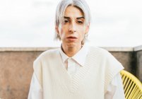 Стильная трансгендерная женщина в модном наряде сидит в кресле на террасе и смотрит в камеру — стоковое фото