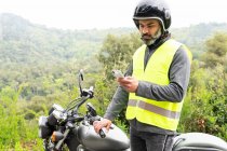 Thoughtful adult Hispanic male biker in protective helmet and vest messaging on mobile phone while standing near broken motorbike near lush green woods — Stock Photo