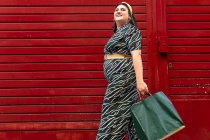 Low angle side view of happy young curvy female in stylish dress with geometric print carrying shopping bags while standing near red striped wall in city — Stock Photo