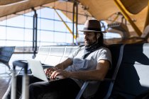 El tipo en el sombrero en el aeropuerto en la sala de espera sentado esperando su vuelo, con auriculares inalámbricos para escuchar música mientras trabaja con su computadora portátil - foto de stock