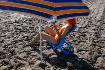 Vue latérale d'une jeune rousse anonyme utilisant le téléphone alors qu'elle était assise sur une chaise à la plage par une journée ensoleillée en été — Photo de stock
