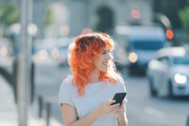Alegre pelirroja mujer en la calle y mensajería en las redes sociales en el teléfono móvil - foto de stock