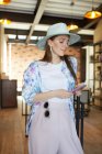 Улыбающаяся молодая женщина в стильной одежде с сотовым телефоном в кафетерии с лампами — стоковое фото