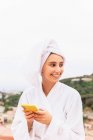 Junge Frau in Bademantel und Handtuch lächelt und surft mit Handy, während sie sich nach der Dusche auf dem Balkon ausruht — Stockfoto