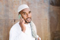 Macho muçulmano alegre em roupas tradicionais sorrindo e navegando celular enquanto está perto de parede rasgada na rua — Fotografia de Stock