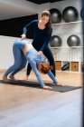 Piena lunghezza di giovane femmina che fa verso il basso Cane posa con l'aiuto di allenatore durante la lezione di yoga in palestra — Foto stock