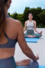 Jeune femme consciente avec les jambes croisées et les yeux fermés pratiquant le yoga contre les cultures partenaire méconnaissable dans la cour — Photo de stock