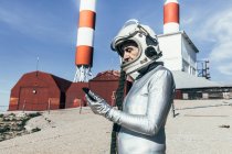 Hombre de edad en traje espacial y datos de navegación casco en el teléfono inteligente, mientras que de pie cerca de edificios industriales con antenas en forma de cohete - foto de stock