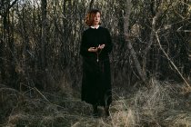 Жінка без емоцій в довгій чорній сукні, що стоїть з хрестом на ланцюжку в осінніх лісах під час святкування Хеллоуїна — стокове фото