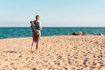 Vista trasera de cuerpo completo de mujer descalza joven irreconocible en ropa deportiva con estera de yoga enrollada mirando a la distancia mientras está de pie en la playa de arena cerca del mar - foto de stock