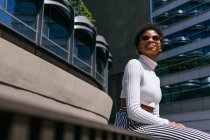 From below positive young African American woman in trendy outfit looking away and enjoying sunlight while sitting on bench on modern city street — Stock Photo