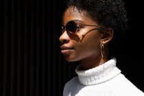 Vista lateral de la joven mujer afroamericana en suéter elegante y gafas de sol mirando hacia otro lado mientras está de pie en la luz del sol brillante contra el fondo negro - foto de stock