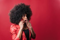 Mujer afroamericana asombrada hablando por teléfono móvil y cubriendo la boca mientras escucha rumores sobre fondo rojo en el estudio - foto de stock