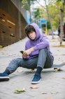 Serious male teenager in casual clothes sitting on skateboard and looking at camera while surfing cellphone on paved sidewalk in city — Stock Photo