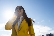 Sorrindo asiático mulher de negócios com casaco amarelo e telefone inteligente andando na rua com edifício no fundo — Fotografia de Stock