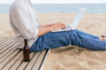 Vista lateral del blogger masculino irreconocible recortado en ropa elegante y sombrero sentado con una botella de cerveza en la playa de arena y escribiendo en el portátil durante las vacaciones de verano en la orilla del mar - foto de stock