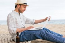 Side view of young male blogger in stylish wear and hat sitting with bottle of beer on sandy beach and typing on laptop during summer holidays on seashore — Stock Photo
