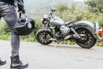 Back view of crop anonymous male biker in jeans and leather gloves holding helmet in hand while standing on asphalt road near parked modern motorcycle — Stock Photo