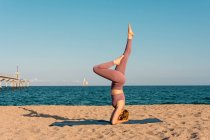 Vista laterale del rilassato bilanciamento femminile a Salamba Sirsasana sul tappeto mentre fai yoga sulla riva del mare — Foto stock