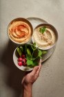 Top view of crop person holding bowls with assorted hummus served on table with fresh cucumbers and radish — Stock Photo