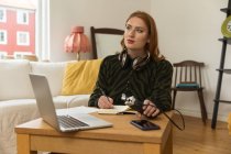 Konzentrierte, nachdenkliche Radiomoderatorin mit Mikrofon und Kopfhörer, die in Notizblock schreibt, während sie sich auf die Aufzeichnung des Podcasts zu Hause vorbereitet — Stockfoto