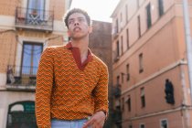 From below young ethnic curly haired guy in stylish colorful striped shirt standing on the street looking at camera pensively — Stock Photo
