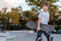 Teenage boy in protective gear standing with skateboard in skate park and looking away — Stock Photo