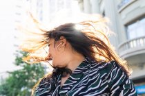 Low angle of cheerful young female in stylish outfit and earrings shaking hair in sunlight on urban street — Stock Photo