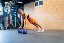 Athlète féminine déterminée dans les vêtements de sport debout dans la pose de planche pendant l'entraînement fonctionnel avec des poids sur le sol dans la salle de gym regardant loin — Photo de stock