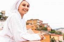 Mujer joven optimista en albornoz y toalla sonriendo y mirando hacia otro lado mientras se relaja en el balcón durante la rutina de cuidado de la piel en fin de semana - foto de stock