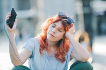Mujer soñadora con el pelo rojo sentada en la calle y escuchando música en los auriculares mientras disfruta de canciones con los ojos cerrados y los brazos extendidos - foto de stock