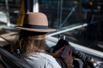 Der Mann mit dem Hut am Flughafen im Wartezimmer, der auf seinen Flug wartet, mit drahtlosen Kopfhörern, um Musik zu hören, während er mit seinem Smartphone chattet, Rückansicht — Stockfoto