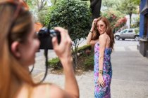 Mujer con cámara fotográfica tomando foto de novia en vestido de verano de pie en la calle y mostrando gesto de encuadre - foto de stock