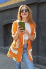 Moda mujer alegre en traje de mezclilla de pie en la calle y navegar por las redes sociales en el teléfono móvil - foto de stock