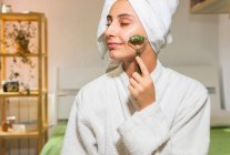 Jovem feliz com toalha na cabeça sorrindo e massageando o rosto com rolo de jade durante a rotina de cuidados com a pele em casa — Fotografia de Stock