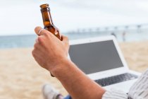 Side view of cropped unrecognizable male sitting with bottle of beer on sandy beach and typing on laptop during summer holidays on seashore — Stock Photo