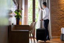 Full body low angle side view of adult man with luggage opening curtain hanging on balcony door in hotel room — Stock Photo