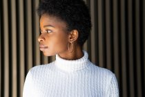 Donna afroamericana indifferente in maglione alla moda guardando lontano contro muro edificio a strisce sulla strada — Foto stock