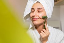 Jovem feliz com toalha na cabeça sorrindo e massageando o rosto com rolo de jade durante a rotina de cuidados com a pele em casa — Fotografia de Stock