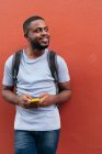 Чорний чоловік з рюкзаком і мобільним телефоном посміхається, спираючись на стіну — стокове фото