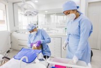 Estomatóloga femenina que trata los dientes de un paciente masculino irreconocible contra su compañero de trabajo en uniforme en el hospital - foto de stock