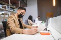 Concentrated ethnic male employee with face mask sitting at table with laptop and reading documents while working in spacious office with blurred colleagues during covid 19 pandemic — Stock Photo